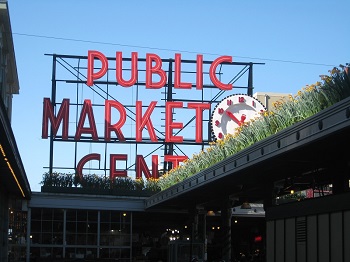 A border of yellow flowers is planted along the edge of a roof of the Pike Place Market in Seattle, Washington, USA.  In the background, a large but partially obscured clock and sign reads 